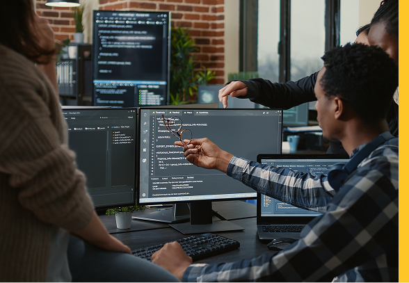 A group of people sitting at a desk with four monitors gesture at code on a screen.