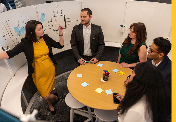 A woman stands in front of a small group around a table and points to a whiteboard with AI written on it.